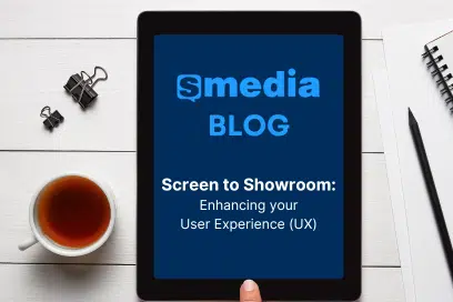 A tablet displaying a blog post titled "Screen to Showroom: Enhancing your User Experience (UX)" from sMedia Blog, placed on a white wooden surface. A hand is touching the screen, and a notebook with a pencil is visible beside it.